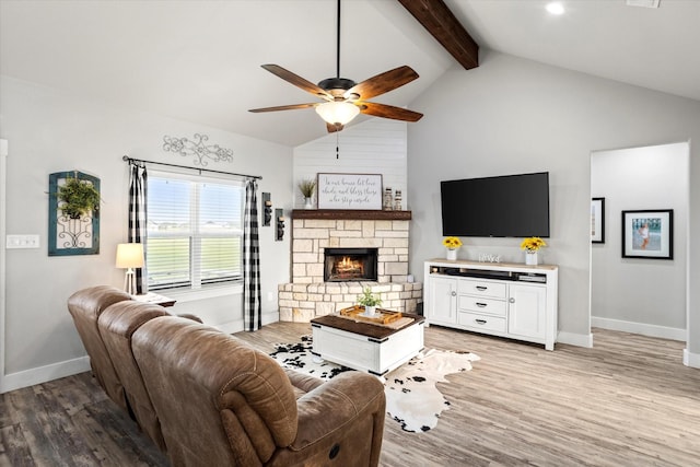 living area featuring beamed ceiling, baseboards, a stone fireplace, a ceiling fan, and light wood finished floors