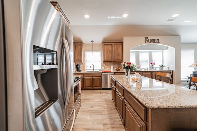 kitchen featuring a healthy amount of sunlight, a sink, a kitchen island, brown cabinetry, and appliances with stainless steel finishes