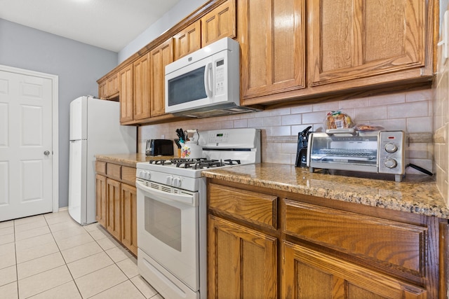kitchen with brown cabinetry, decorative backsplash, light tile patterned flooring, white appliances, and stone counters