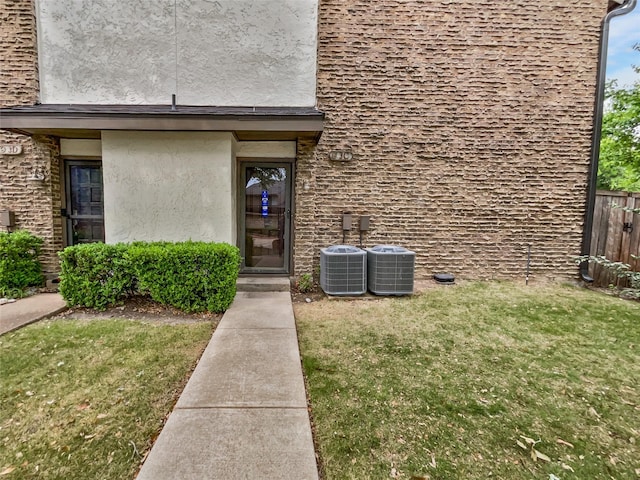 view of exterior entry with central air condition unit, fence, a yard, and stucco siding