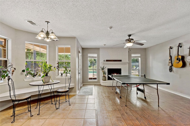 playroom with ceiling fan with notable chandelier, a brick fireplace, visible vents, and a healthy amount of sunlight