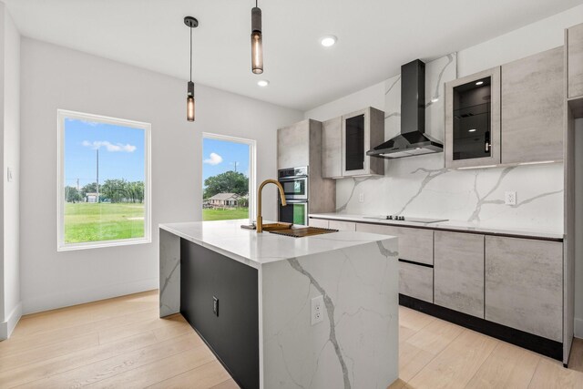 1926 Pueblo St, Dallas, TX 75212 - kitchen with light stone countertops, a kitchen island with sink, light wood-style flooring, wall chimney exhaust hood, and decorative backsplash