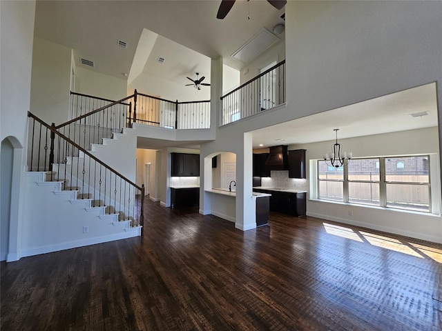 unfurnished living room featuring a high ceiling, stairs, dark wood-style floors, and ceiling fan with notable chandelier