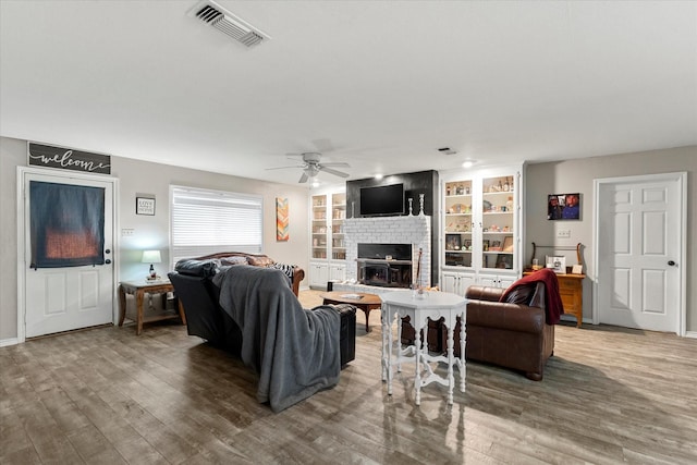 living room featuring wood finished floors, visible vents, a ceiling fan, a fireplace, and built in shelves