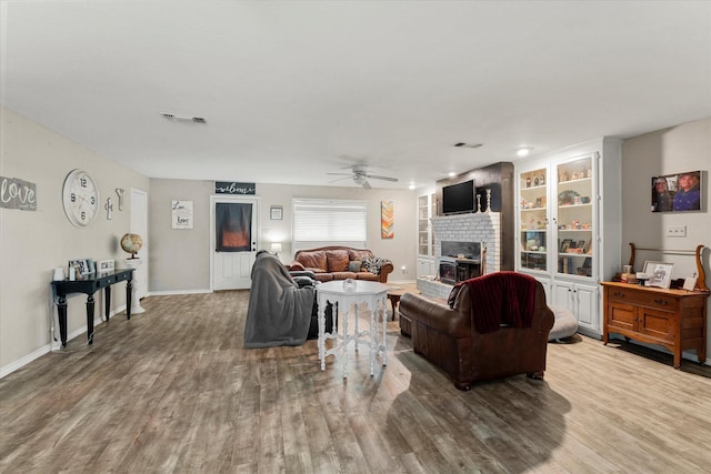 living area featuring baseboards, visible vents, wood finished floors, a brick fireplace, and ceiling fan