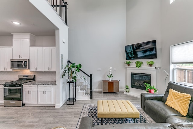 living area featuring stairway, a glass covered fireplace, light wood-style flooring, a high ceiling, and recessed lighting