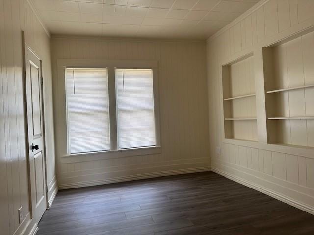 spare room featuring a decorative wall, built in shelves, baseboards, ornamental molding, and dark wood-type flooring