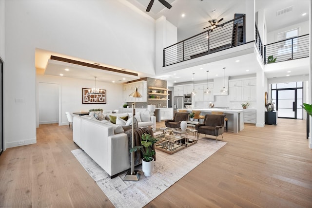 living area featuring a high ceiling, light wood finished floors, visible vents, baseboards, and ceiling fan with notable chandelier