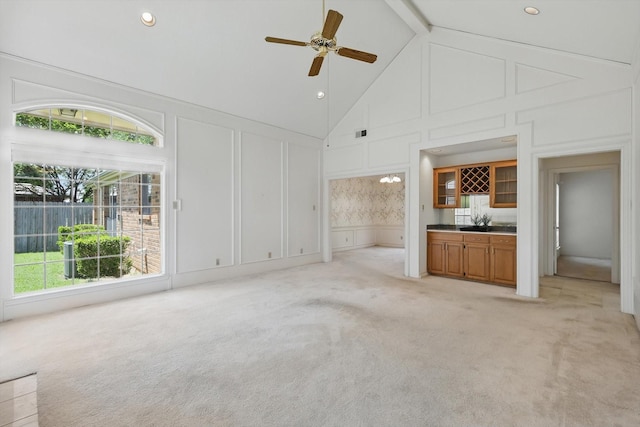 unfurnished living room with high vaulted ceiling, a decorative wall, a ceiling fan, and light carpet