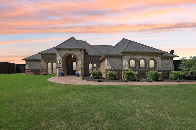 french country inspired facade featuring a lawn, roof with shingles, fence, and brick siding