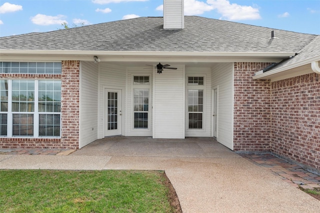 view of exterior entry featuring a chimney, a shingled roof, a patio, brick siding, and ceiling fan