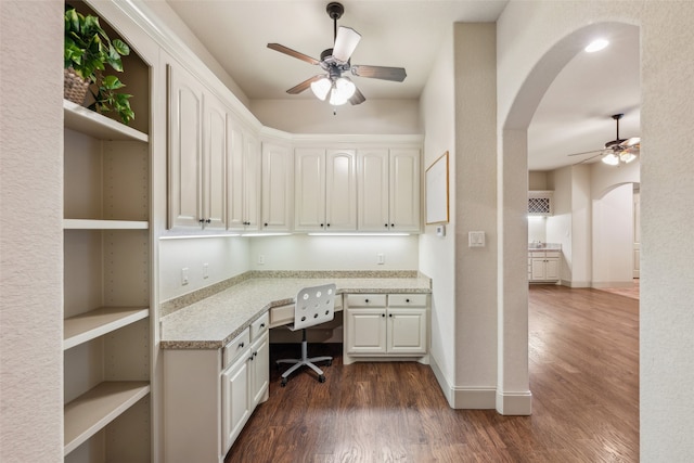 unfurnished office featuring arched walkways, a ceiling fan, dark wood-style floors, and built in desk