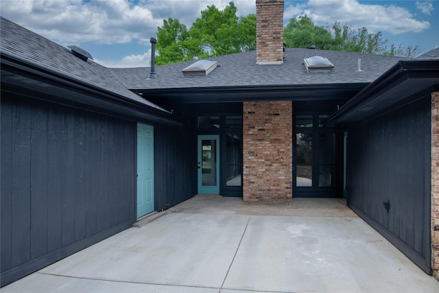 property entrance with brick siding, a patio area, a shingled roof, and a chimney