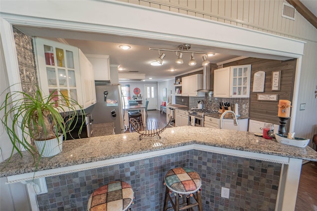 kitchen featuring wall chimney exhaust hood, stainless steel appliances, visible vents, backsplash, and glass insert cabinets