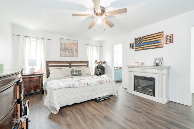 bedroom featuring wood finished floors, connected bathroom, baseboards, a glass covered fireplace, and ceiling fan