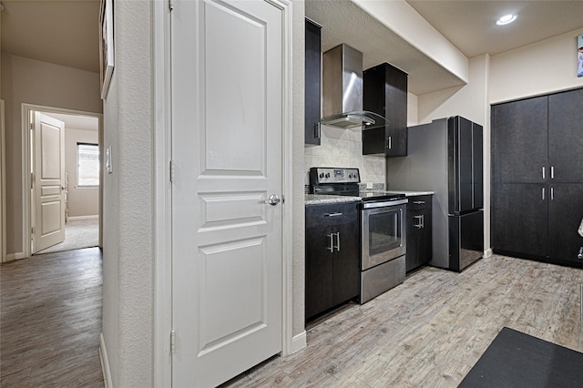 kitchen with wall chimney range hood, tasteful backsplash, stainless steel appliances, light wood-style flooring, and dark cabinets
