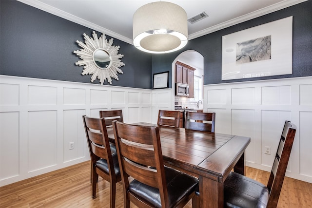 dining area with arched walkways, light wood-type flooring, visible vents, crown molding, and a decorative wall