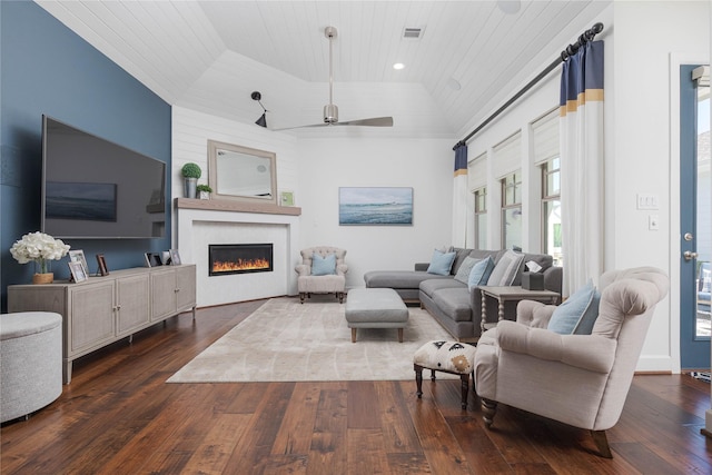 living area featuring a warm lit fireplace, ceiling fan, a raised ceiling, wooden ceiling, and dark wood-style flooring