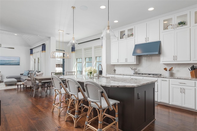 kitchen featuring open floor plan, stainless steel gas stovetop, light stone countertops, under cabinet range hood, and an island with sink