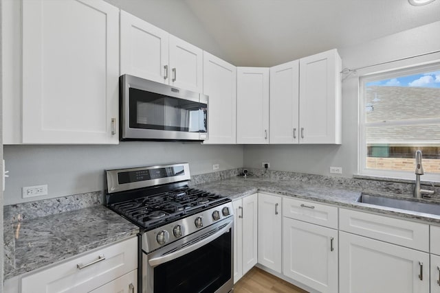 kitchen with vaulted ceiling, light stone counters, a sink, white cabinets, and appliances with stainless steel finishes