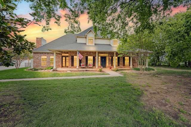 view of front of home featuring a chimney, a shingled roof, a porch, a lawn, and brick siding