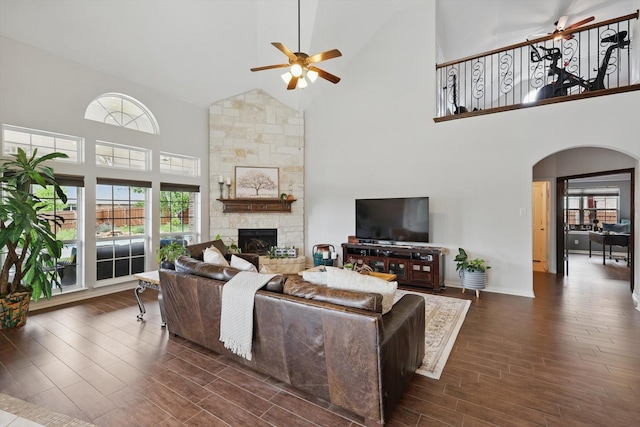 living area with a stone fireplace, dark wood-type flooring, arched walkways, high vaulted ceiling, and ceiling fan