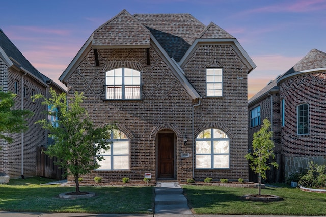 french provincial home with roof with shingles, brick siding, fence, and a yard