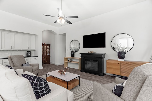 living area featuring arched walkways, a ceiling fan, a glass covered fireplace, and light wood-style flooring