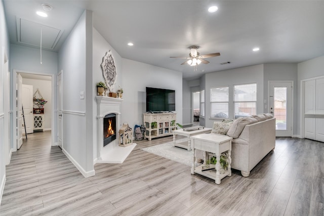 living area with attic access, ceiling fan, a lit fireplace, and light wood-style flooring