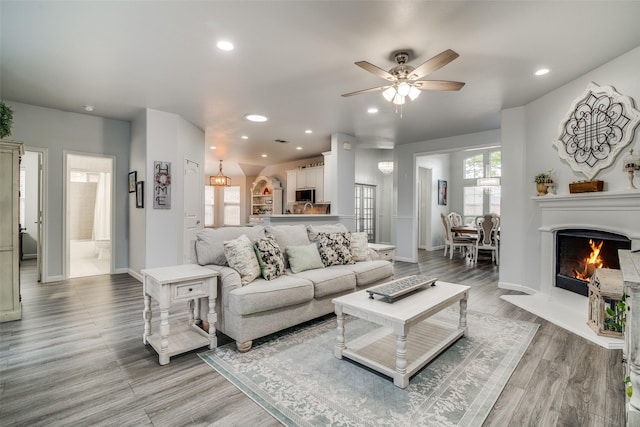 living area featuring light wood-style flooring, baseboards, recessed lighting, a warm lit fireplace, and ceiling fan