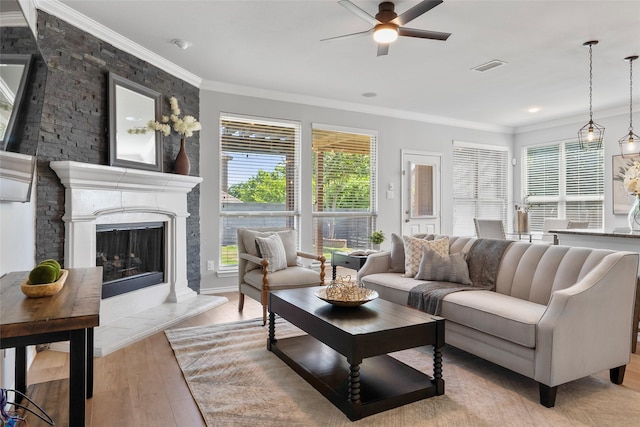 living area with light wood-style floors, a fireplace with raised hearth, a ceiling fan, visible vents, and ornamental molding