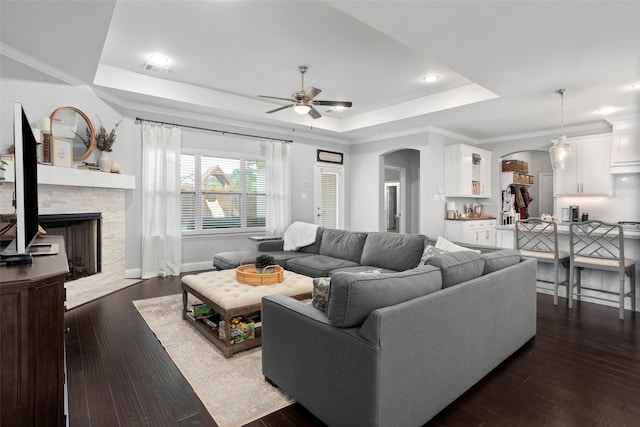 living room with a tray ceiling, arched walkways, visible vents, a ceiling fan, and a stone fireplace