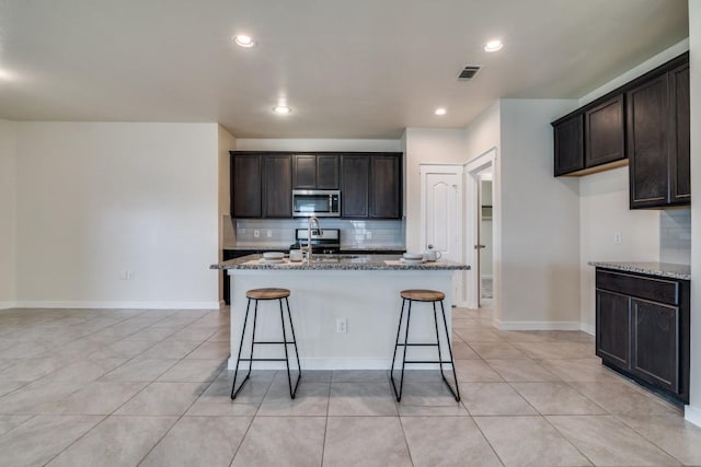 kitchen with stainless steel microwave, a kitchen island with sink, visible vents, backsplash, and stone countertops