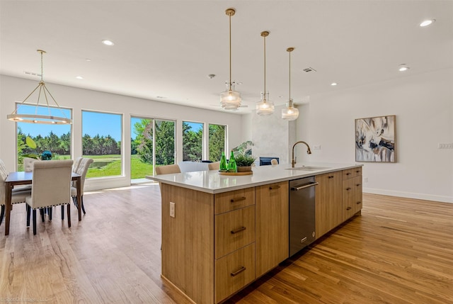 kitchen with light wood finished floors, stainless steel dishwasher, a sink, a center island with sink, and light countertops