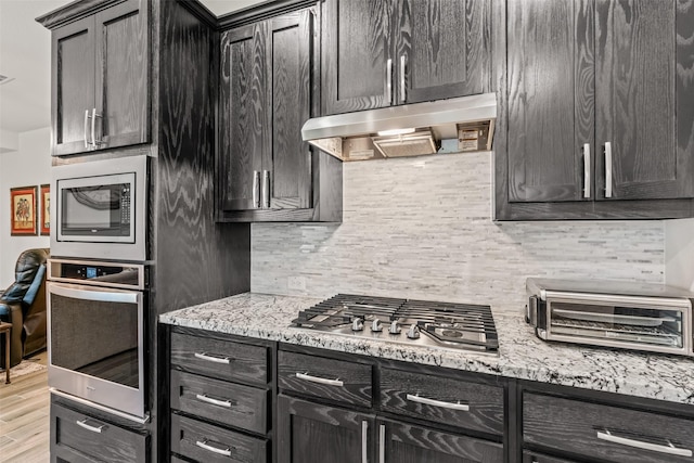 kitchen featuring light stone counters, stainless steel appliances, light wood finished floors, backsplash, and under cabinet range hood