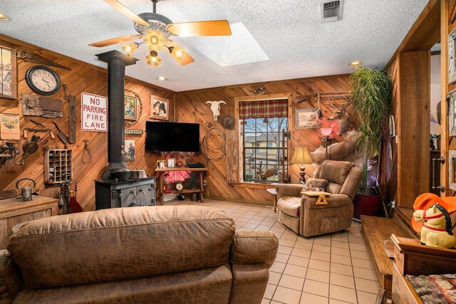 living area featuring wooden walls, tile patterned flooring, a wood stove, a ceiling fan, and visible vents