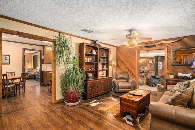 living area featuring ceiling fan, a textured ceiling, a wood stove, dark wood finished floors, and wooden walls