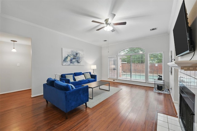 living room featuring a tiled fireplace, ceiling fan, visible vents, ornamental molding, and wood finished floors