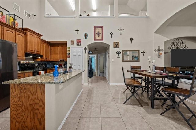 kitchen with a towering ceiling, under cabinet range hood, arched walkways, light tile patterned floors, and black appliances