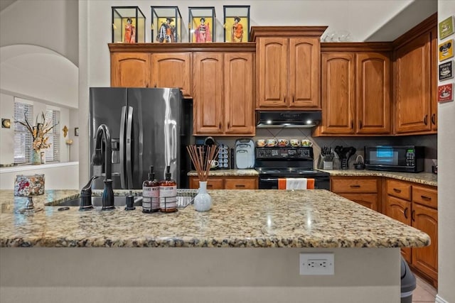 kitchen with under cabinet range hood, black appliances, light stone countertops, and brown cabinets