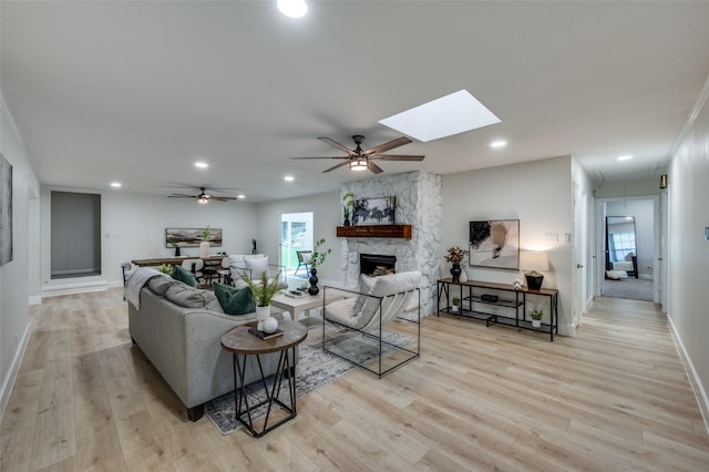 living area featuring a stone fireplace, light wood-type flooring, a ceiling fan, a skylight, and baseboards