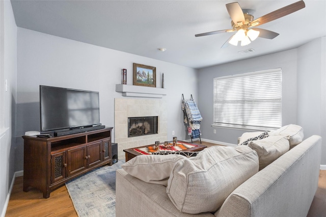 living room with ceiling fan, light wood finished floors, a tiled fireplace, baseboards, and visible vents