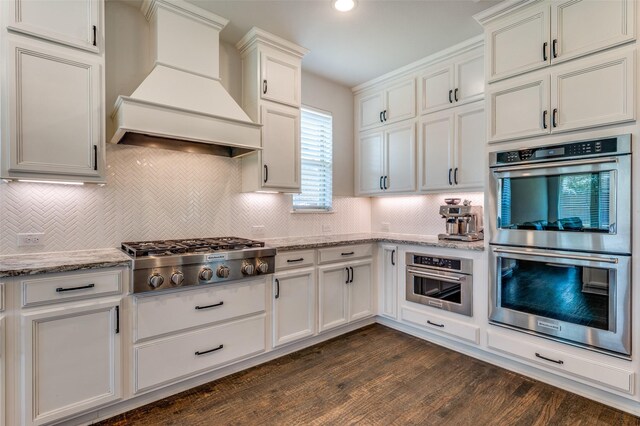 kitchen featuring light stone counters, decorative backsplash, appliances with stainless steel finishes, white cabinetry, and custom range hood
