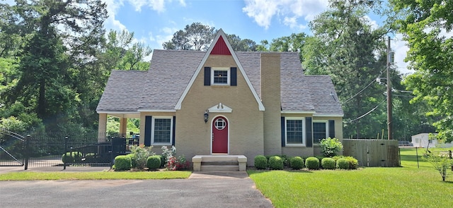tudor-style house with brick siding, fence, roof with shingles, and a front lawn