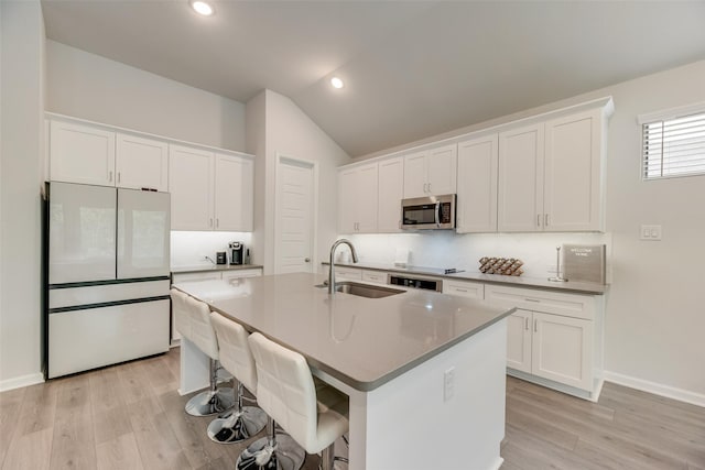 kitchen featuring vaulted ceiling, a sink, an island with sink, stainless steel microwave, and refrigerator with glass door