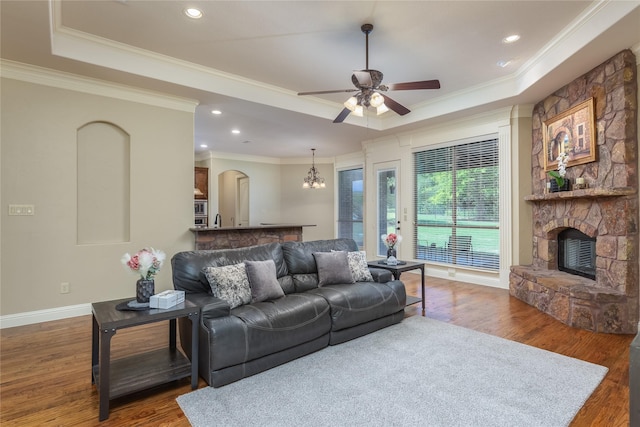 living area with wood finished floors, ceiling fan with notable chandelier, a tray ceiling, a fireplace, and ornamental molding