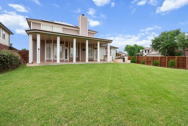 back of house with a fenced backyard, a chimney, a lawn, roof mounted solar panels, and a patio