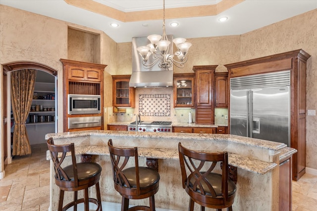 kitchen with brown cabinetry, wall chimney range hood, built in appliances, and glass insert cabinets