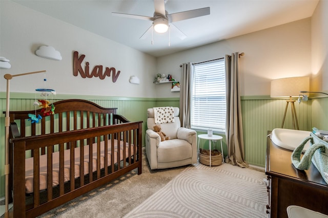 carpeted bedroom featuring wainscoting, a ceiling fan, and a crib