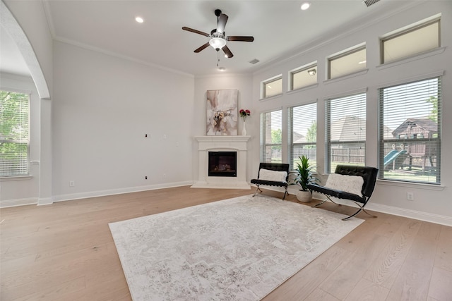 sitting room with light wood-style floors, a fireplace with raised hearth, ceiling fan, and ornamental molding